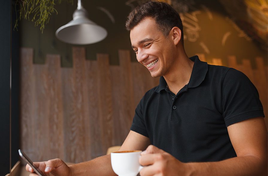 A man smiling while using a phone and holding a cup of coffee