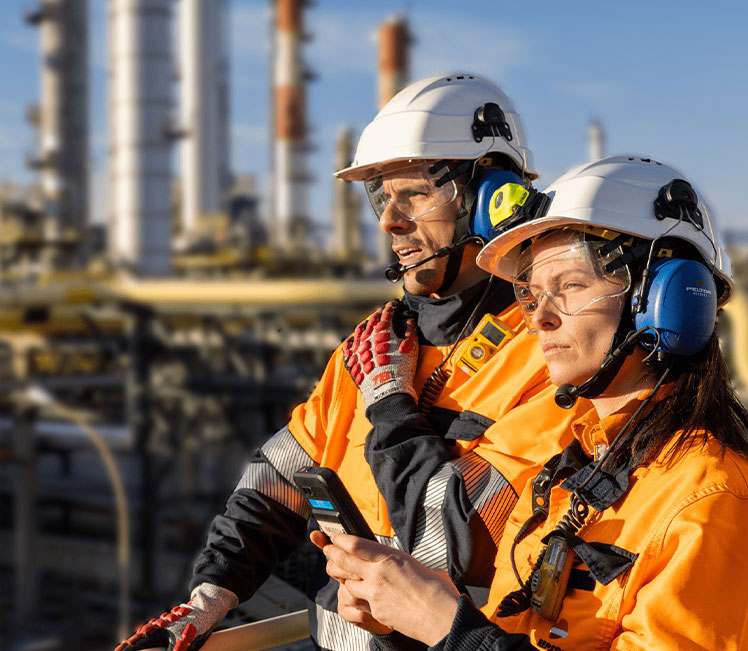 La imagen muestra dos operarios vestidos con ropa de seguridad industrial —casco, chaqueta reflectante de color naranja y chaleco— caminando entre estructuras metálicas y tuberías propias de una planta industrial. Al fondo se observan columnas y conductos elevados, así como elementos estructurales como escalas y plataformas metálicas. Todo indica que es un entorno industrial complejo y funcional, probablemente asociado a una refinería o planta química.