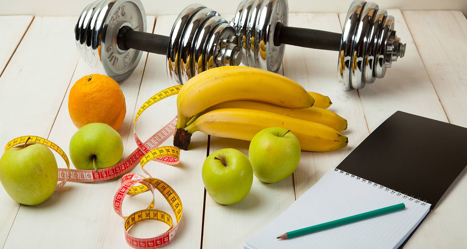 Image of weights together with fruit, a notebook, and a measuring tape.