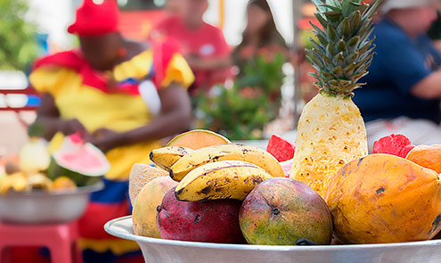 Detalle de un plato de frutas y al fondo una mujer ataviada con los colores de la bandera colombiana 